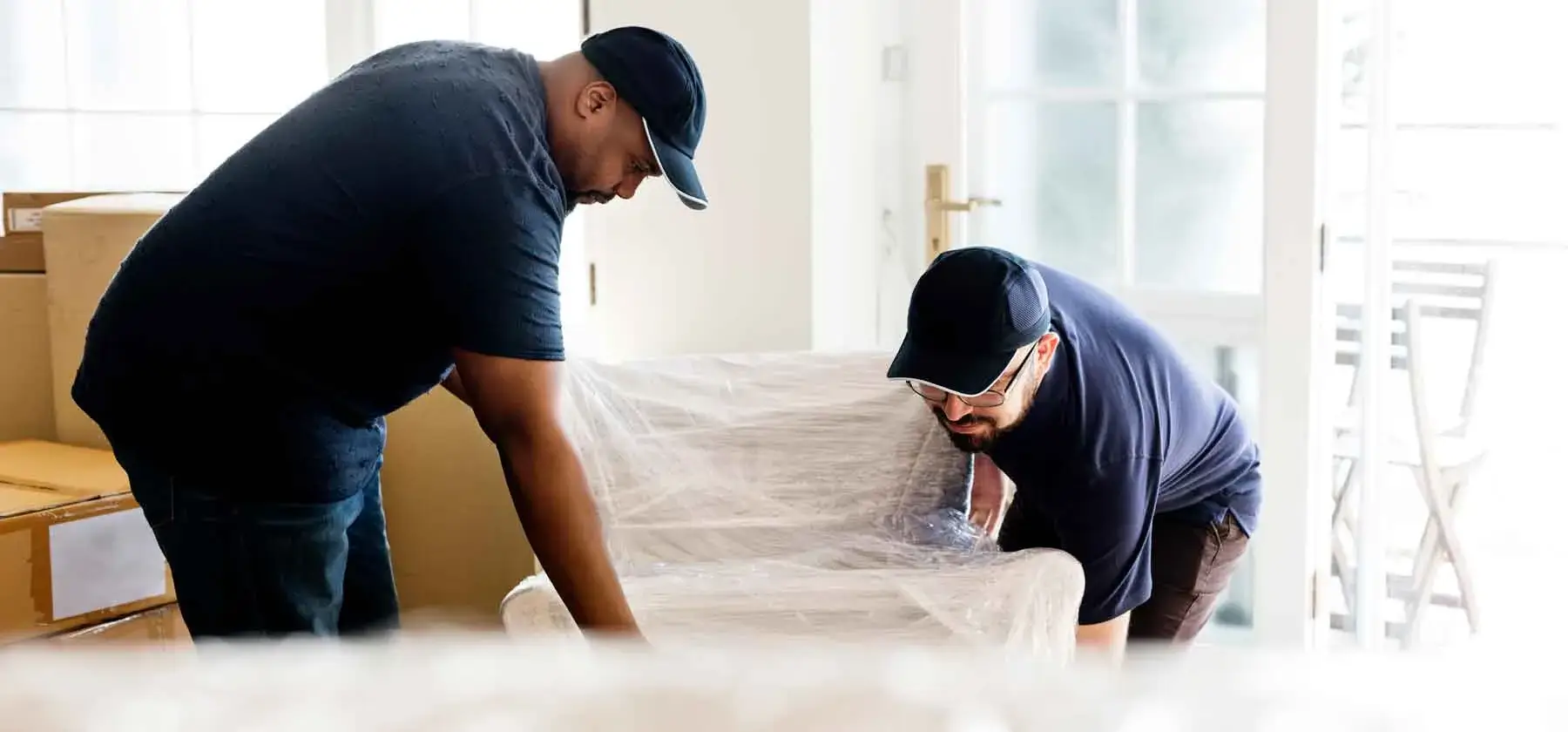 Two men moving wrapped furniture indoors.